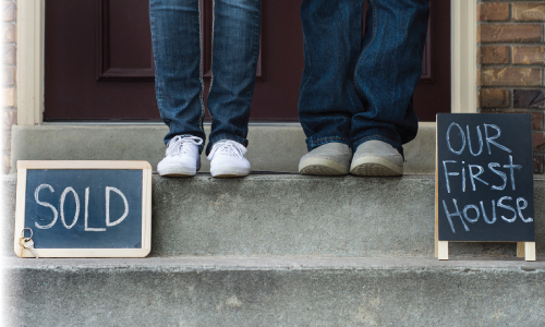 Newhome2 A couple standing on porch of their new house with a sign that says our first house