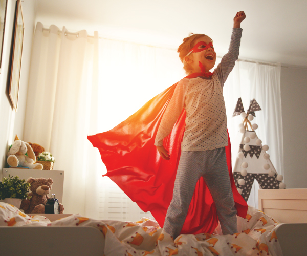 young child wearing a red cape and jumping on the bed