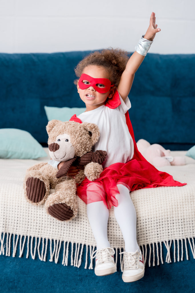 Adorable little african american child in superhero costume sitting on bed with teddy bear